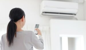 On voit une femme de dos, avec une télécommande dans les mains, face à une climatisation.