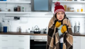 jeune fille dans une maison avec un plaid sur ses épaules, un bonnet et des gants.